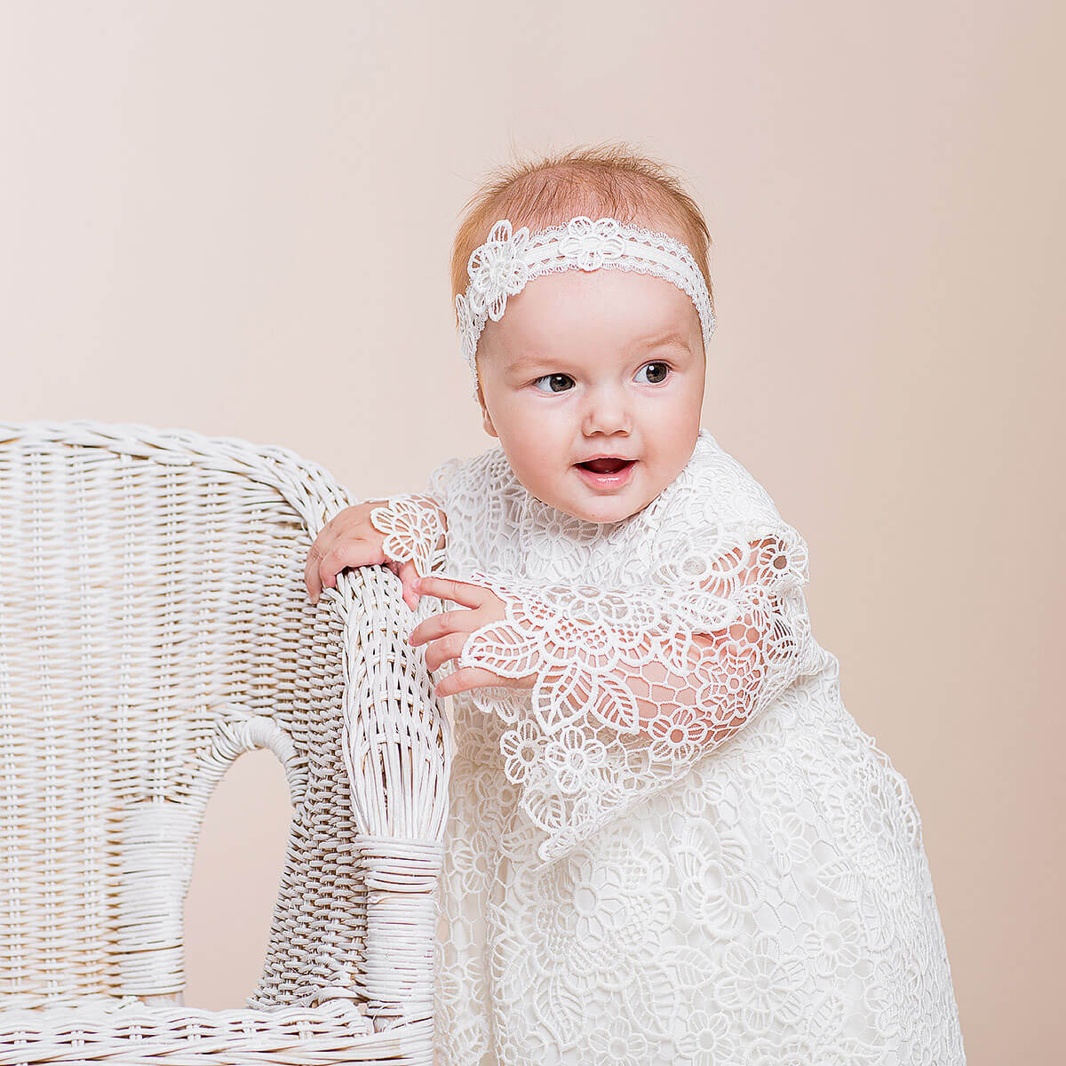 Baby in a Baby Baptism Dress and headband sitting on a wicker chair against a beige background