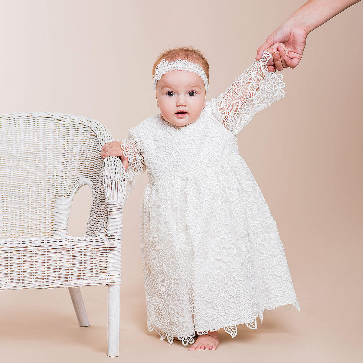 Baby in a Baby Baptism Dress standing next to a wicker chair on a beige background