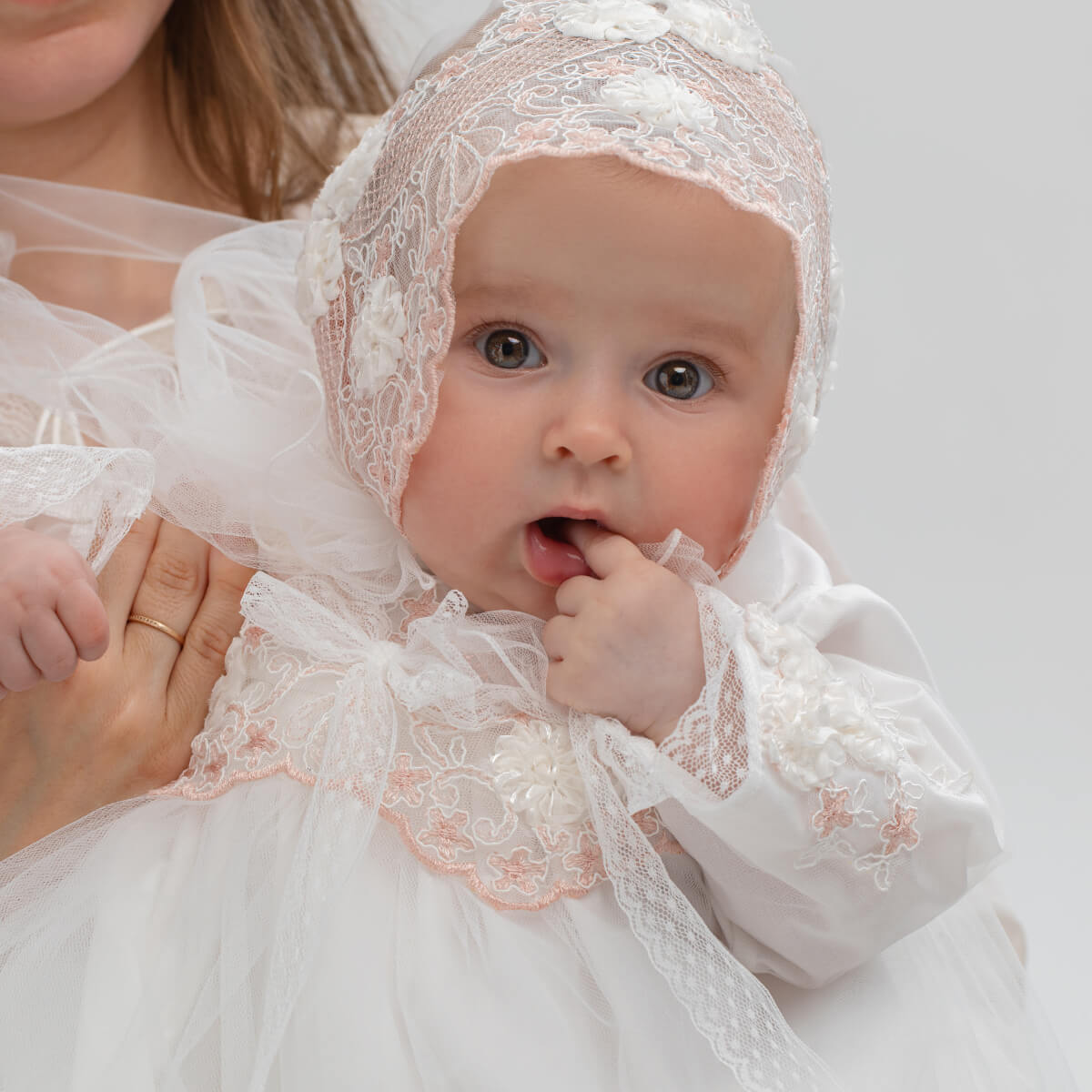 Baby in a flower lace baptism bonnet being held by an adult against a plain background