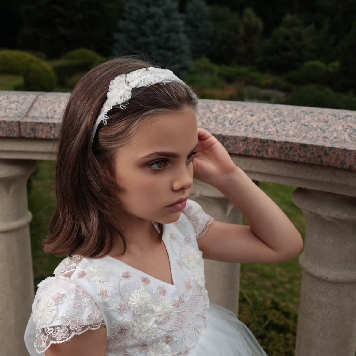 Young girl in a flower girl dress with train with a floral headband, standing outdoors.