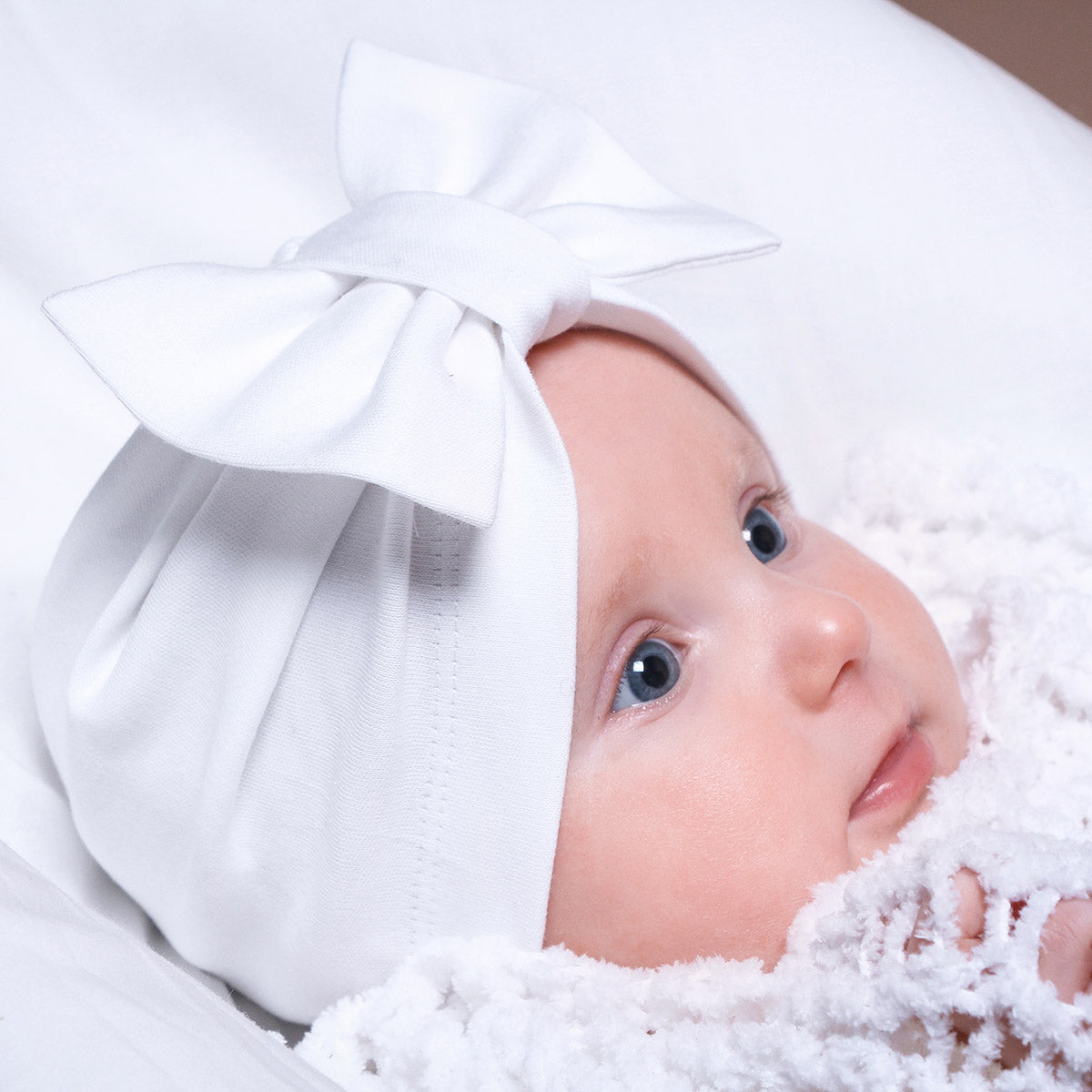 Baby wearing a white baby beanie with a large bow, surrounded by soft white fabric.
