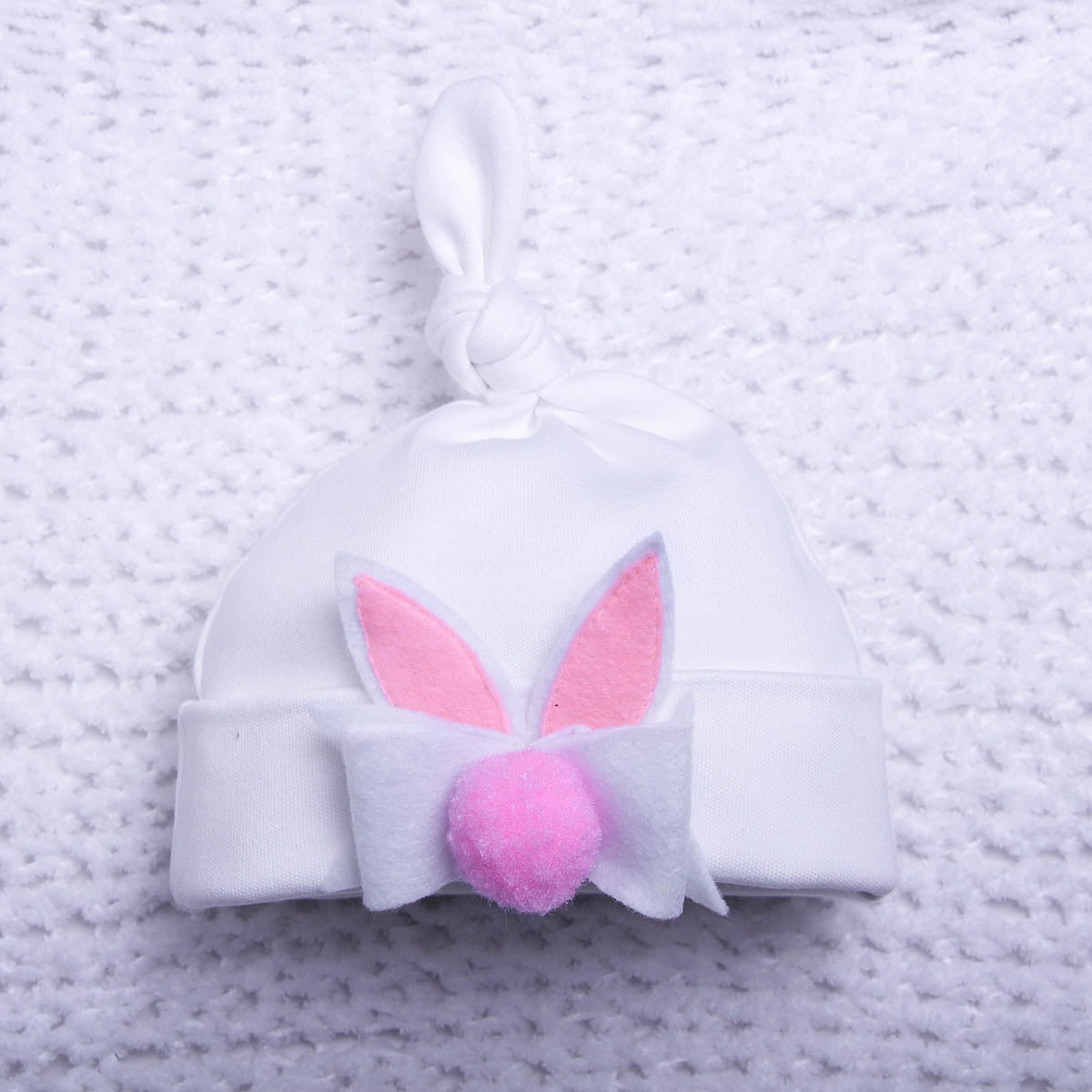 White baby hat with pink bunny ears and a pom-pom on a textured gray background