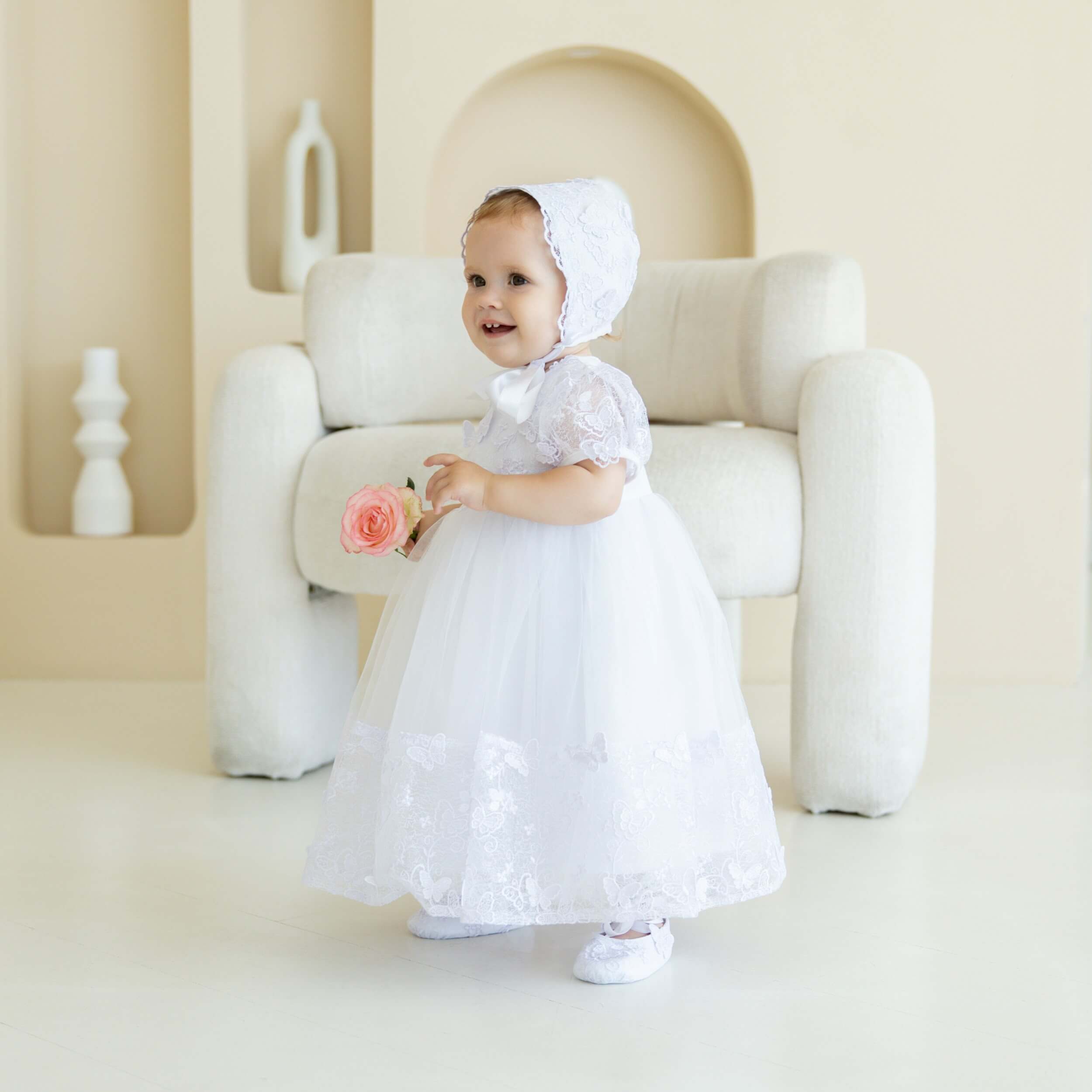 Baby in a white dress and lace baptism bonnet holding a flower in a minimalistic room.