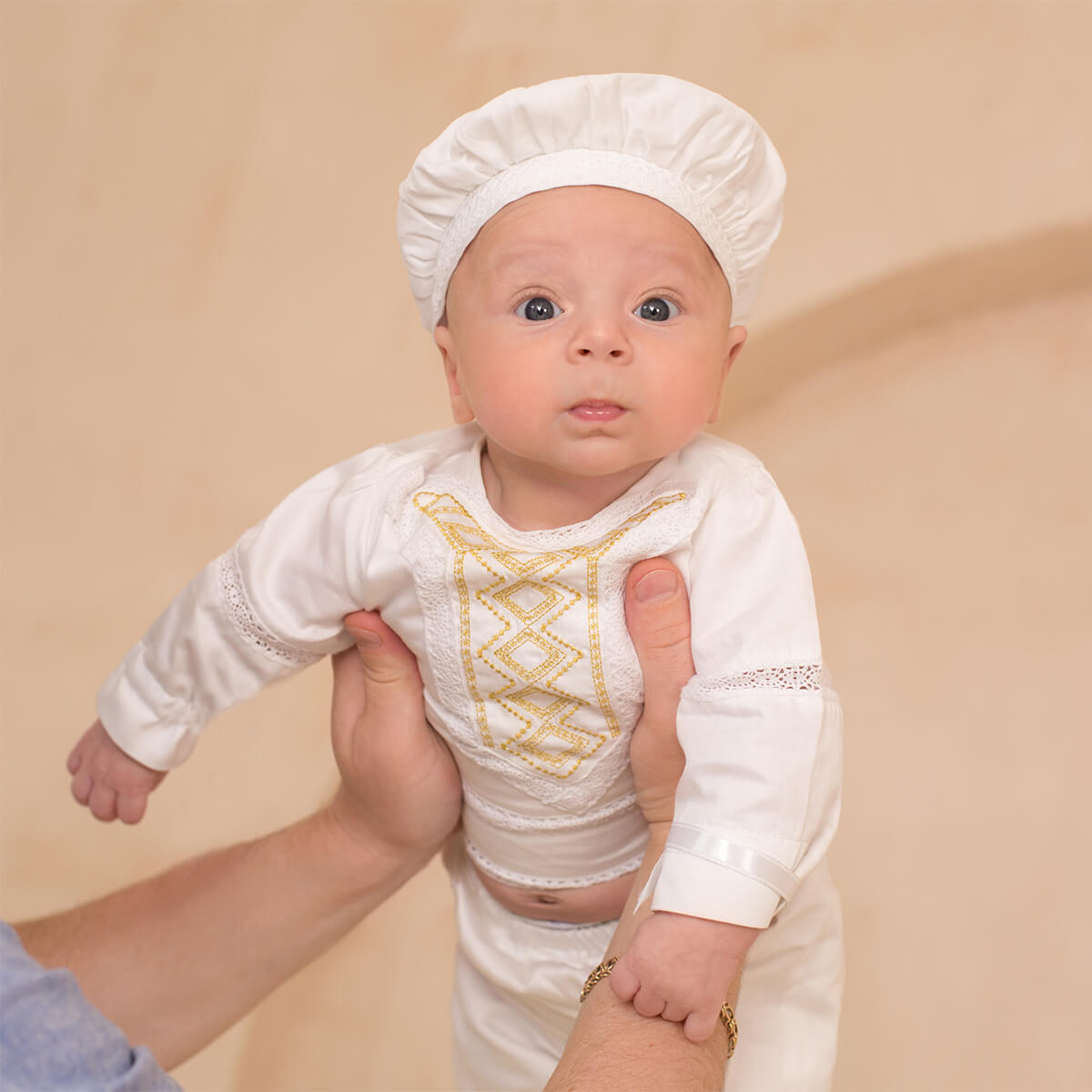 Baby in a Baby Boy Baptism Suit with gold patterns and a matching hat on a beige background