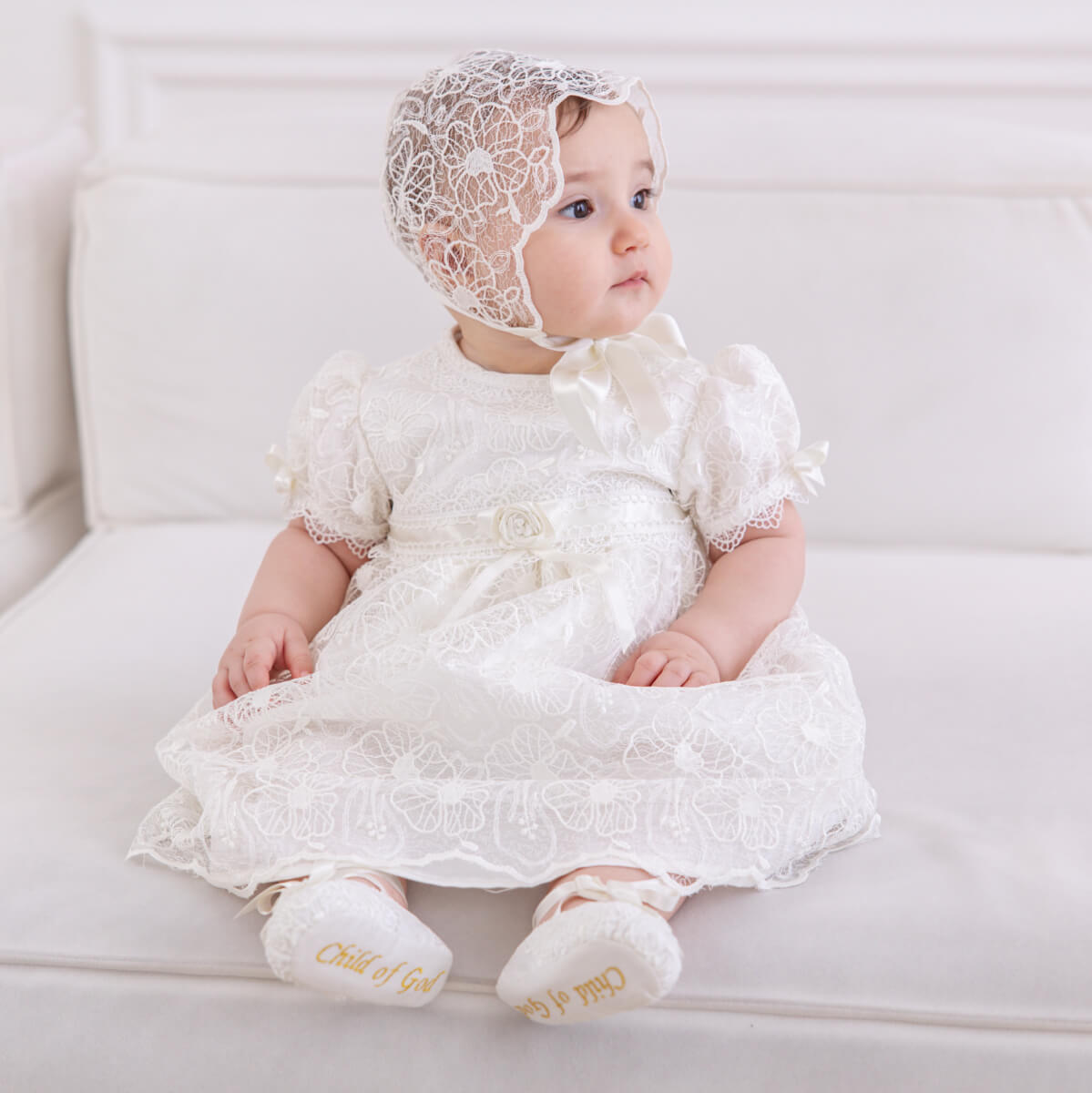 Baby in a Lace Baptism Dress and bonnet sitting on a white couch.