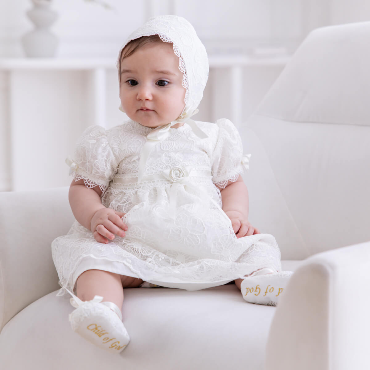 Baby in a Lace Baptism Dress and bonnet sitting on a white couch.