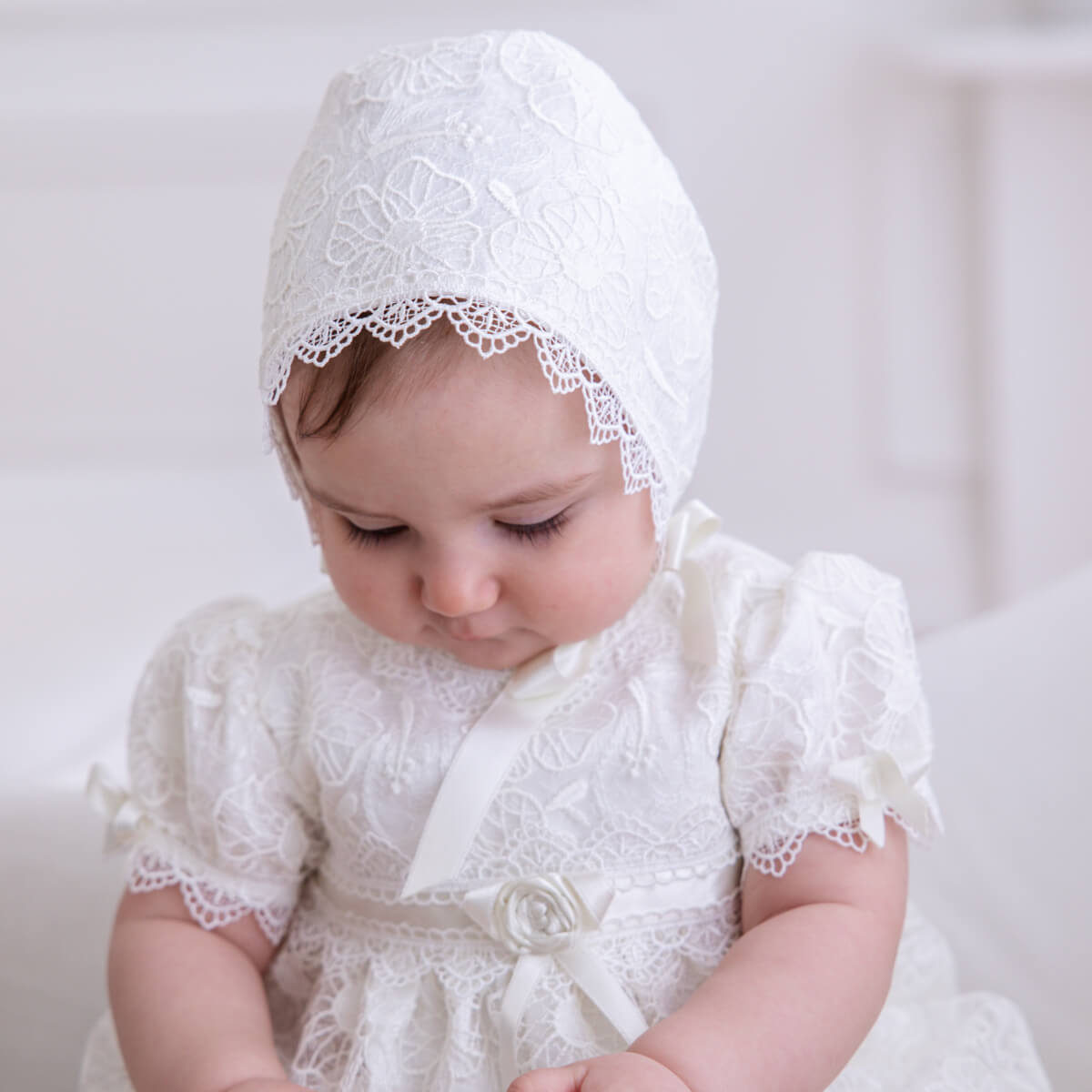 Baby wearing a Lace Baptism Dress and bonnet against a plain background