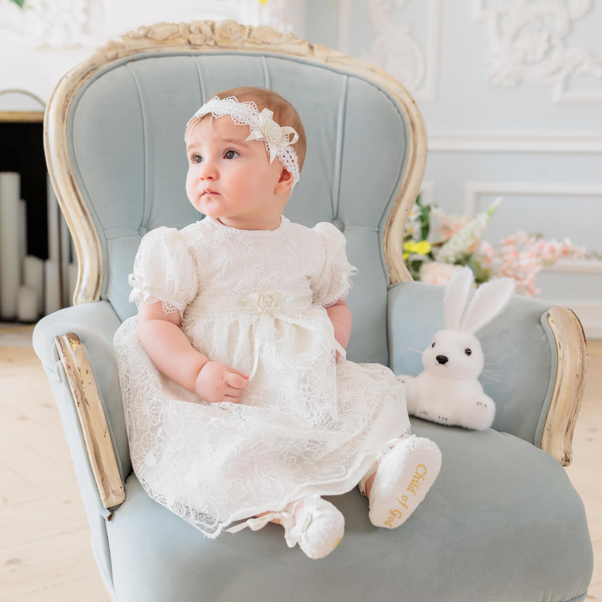 Baby in a Lace Baptism Dress sitting on a light blue chair with a stuffed bunny toy.