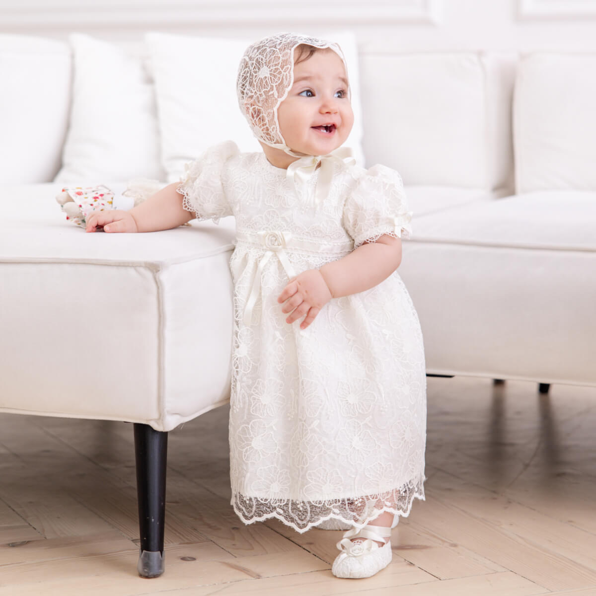 Baby in a Lace Baptism Dress and bonnet sitting on a white couch.