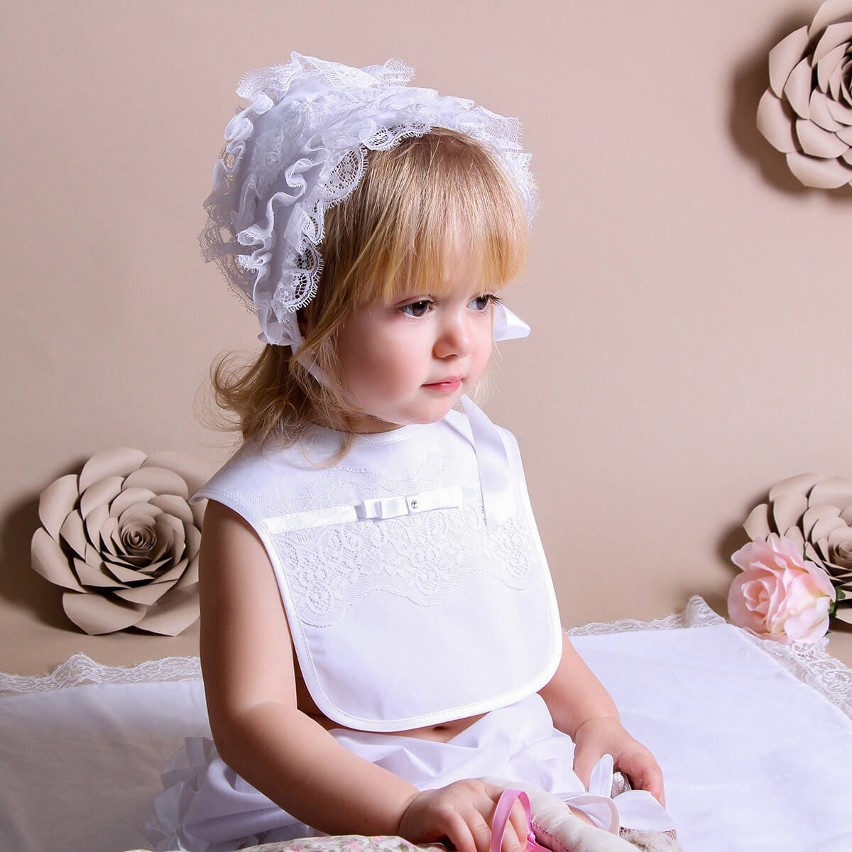Young girl in a white dress with a Baptism Bib sitting on a beige couch.