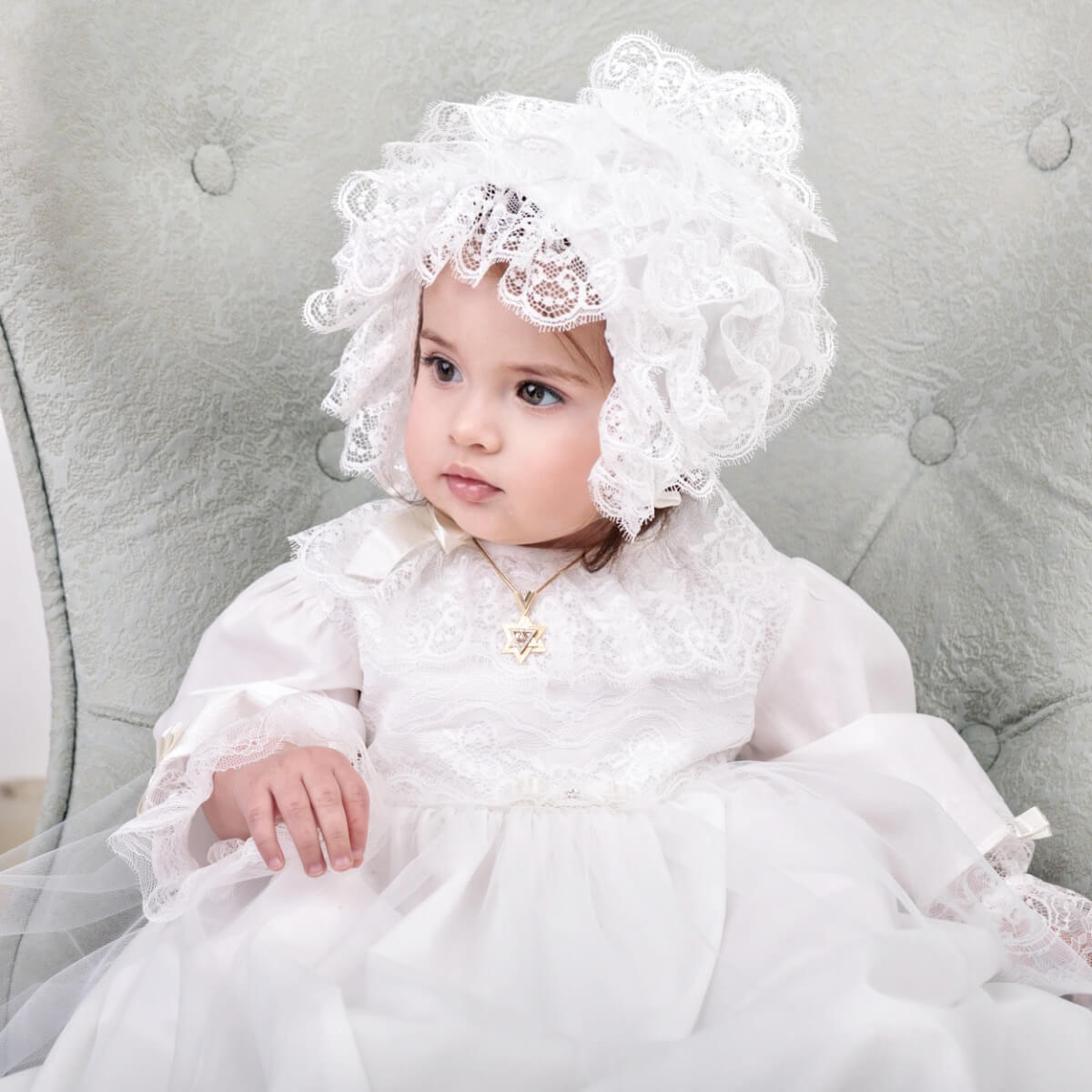 Baby in a white lace dress and Lace Baptism Bonnet sitting on a gray cushion.