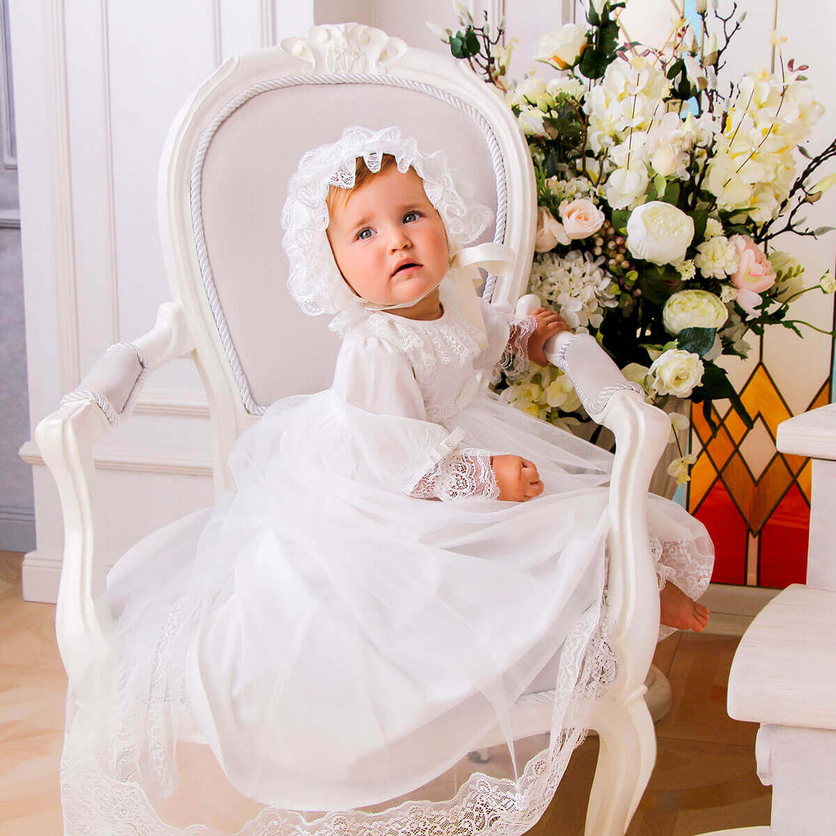 Baby in a Christening Gown for Girls sitting on a white chair with flowers in the background