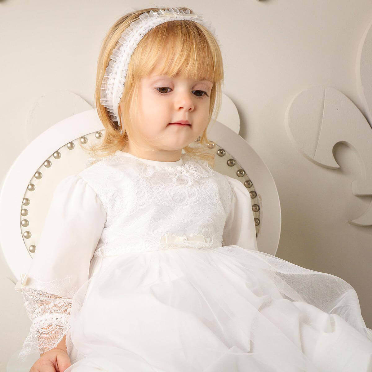 Young girl in a Christening Gown for Girls sitting on a decorative chair.