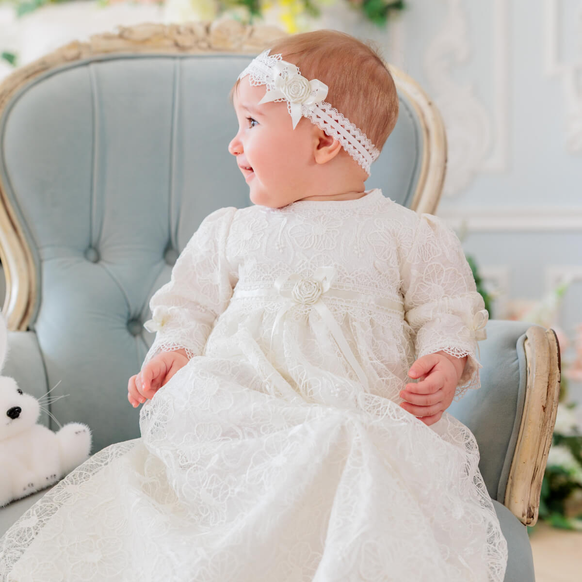 Baby in a Christening Gown for Girls sitting on a light blue chair with a white teddy bear.