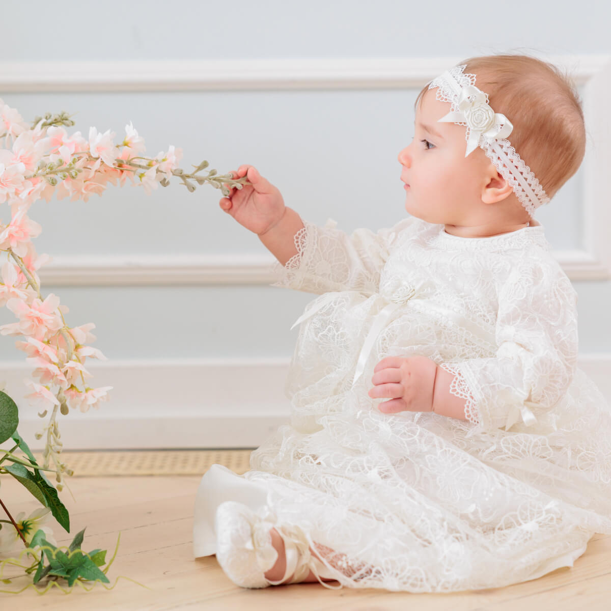 Baby in a Christening Gown for Girls sitting on a light wooden floor with a floral wreath.