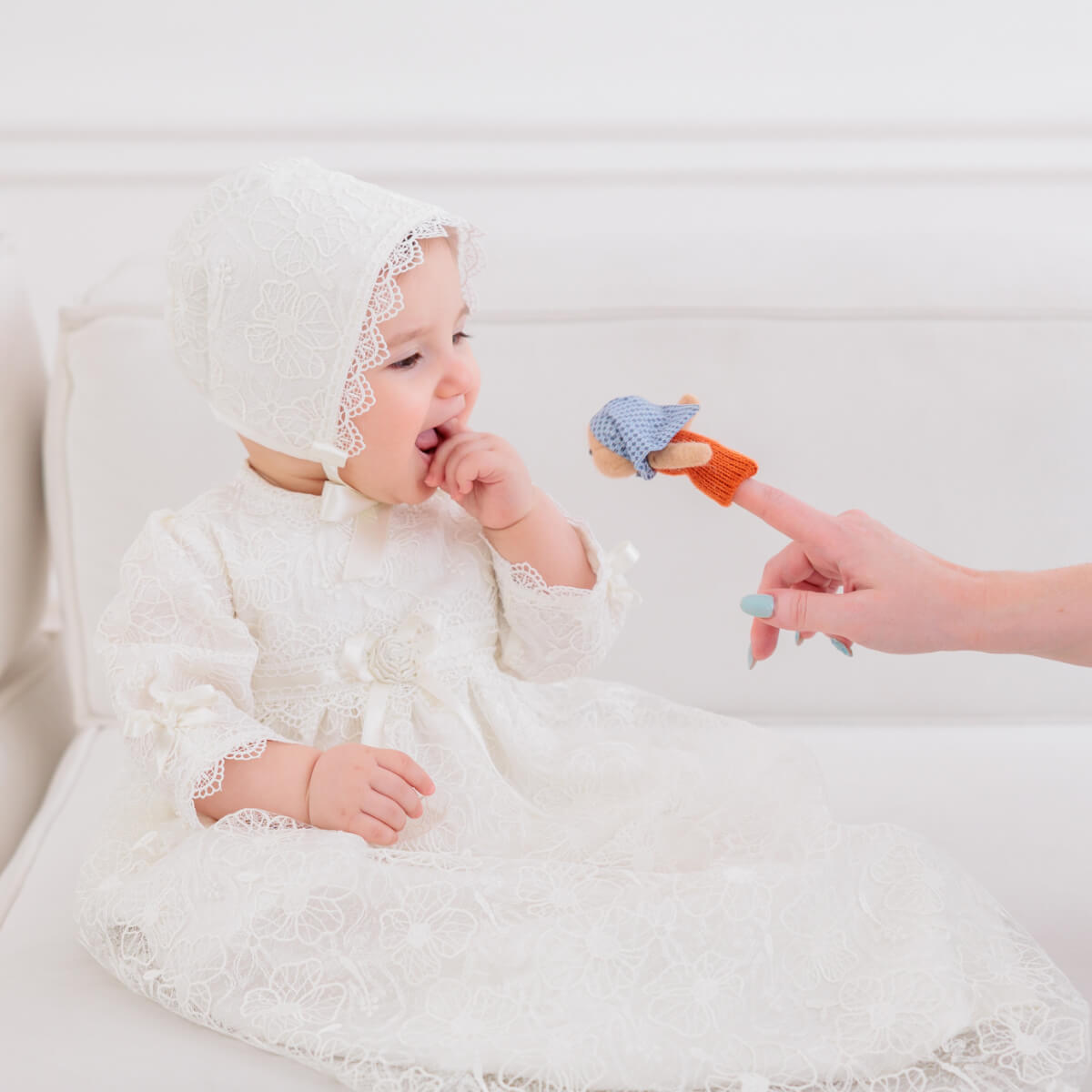 Baby in a white Christening Gown for Girls being offered a small toy by an adult hand.