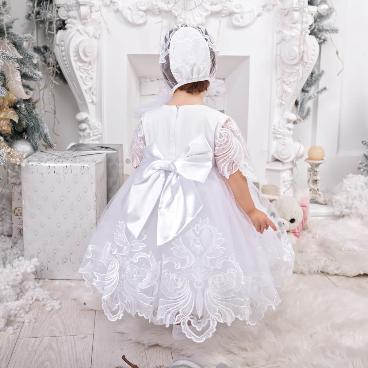 Child in a White Baptism Dress standing in a decorated room with Christmas trees.