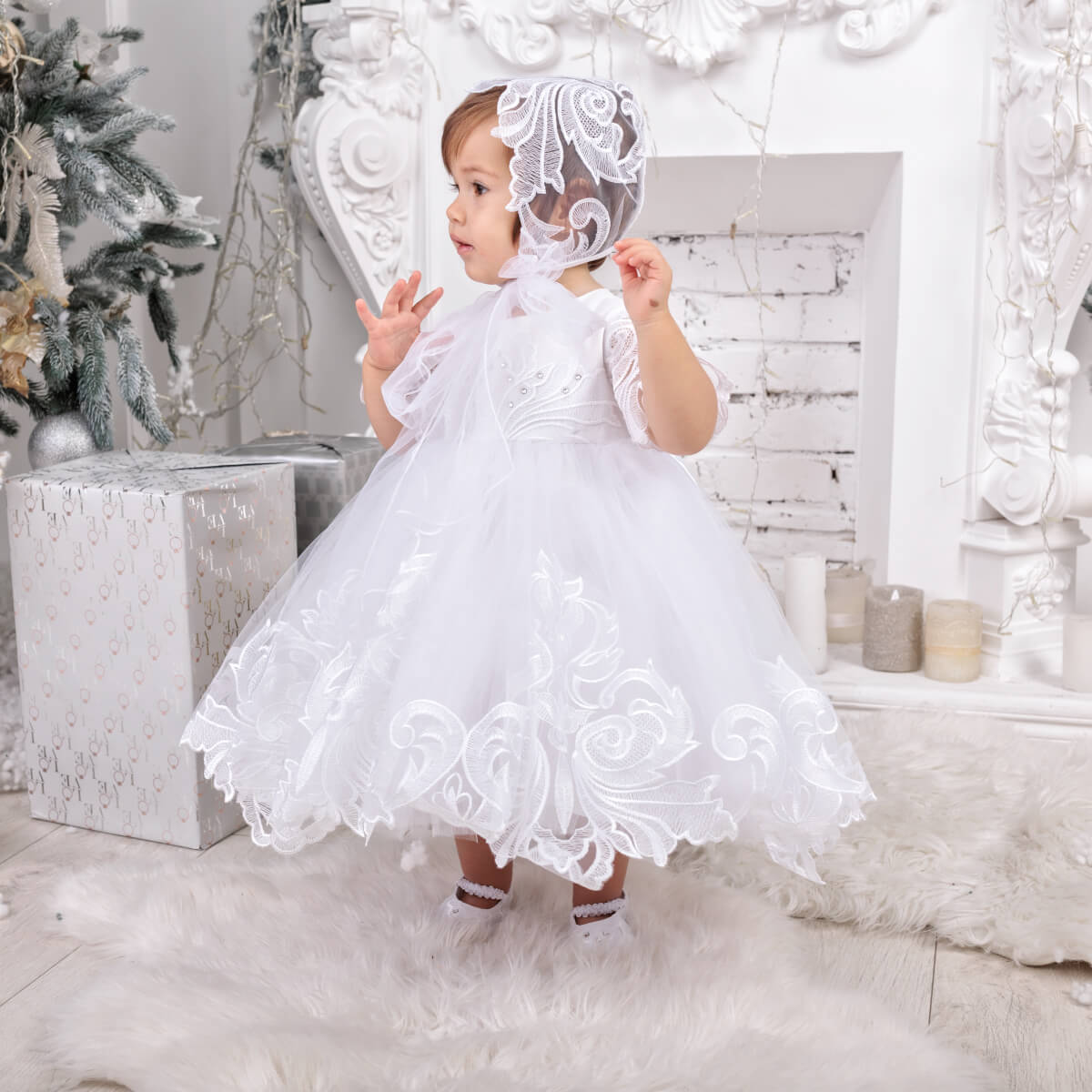 Baby in a White Baptism Dress and bonnet standing in a decorated room.