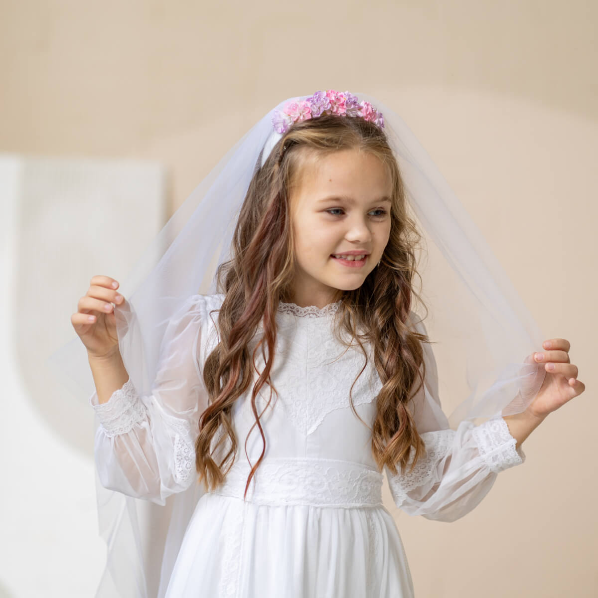 Young girl in a Lace Communion Dress with a veil and floral headband against a beige background