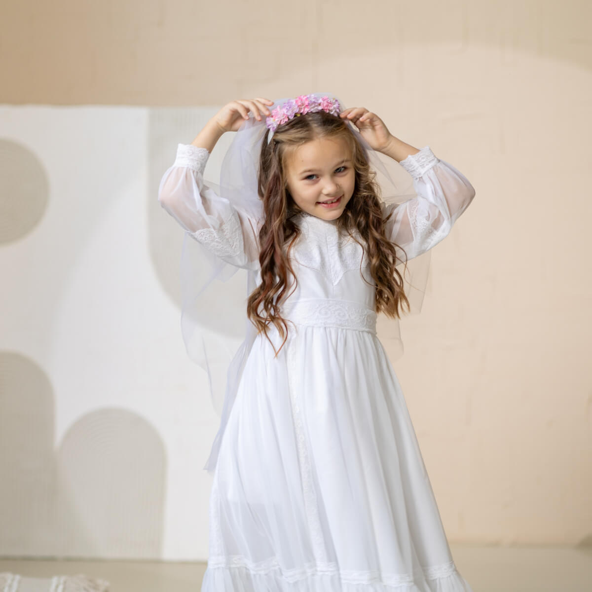 Young girl in a white dress with sheer sleeves and a floral headband against a beige background
