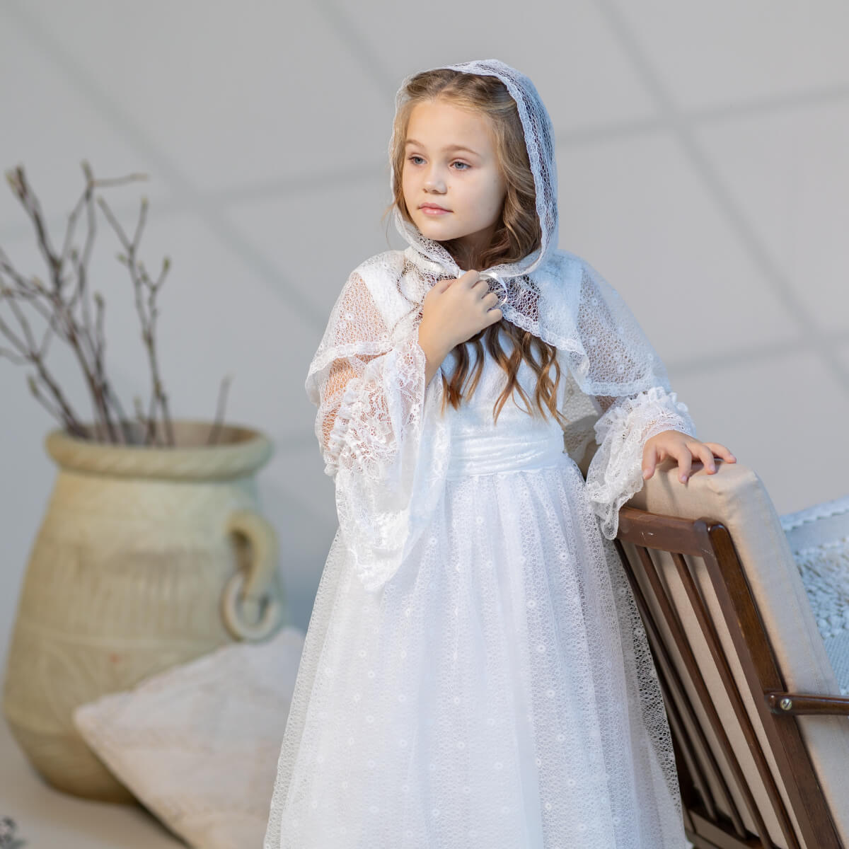 Young girl in a white lace dress and First Communion Mantilla Veil standing indoors with a neutral background