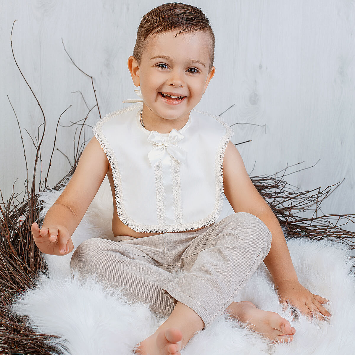 Child wearing a Baptism Bib and beige pants sitting on a white surface with a neutral background