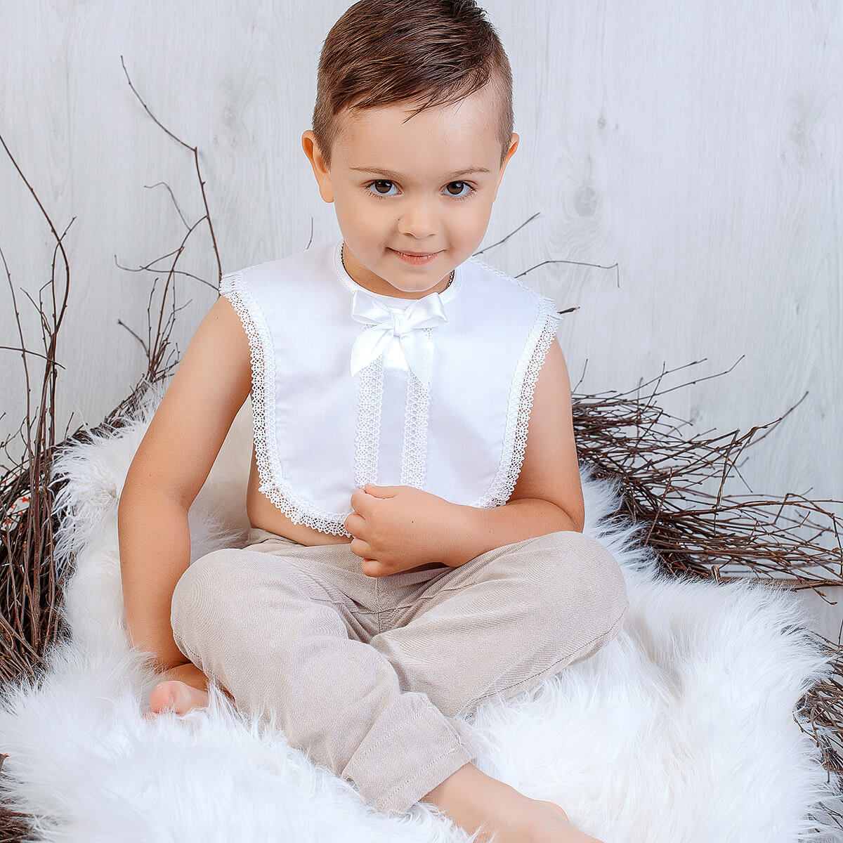 Child wearing a Baptism Bib and beige pants sitting on a fluffy white surface with a neutral background.