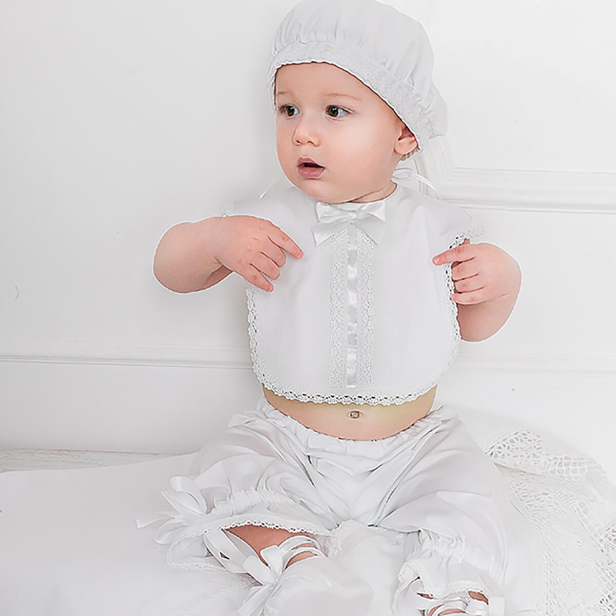 Baby in a white outfit with a Baptism Bib and matching hat on a light background