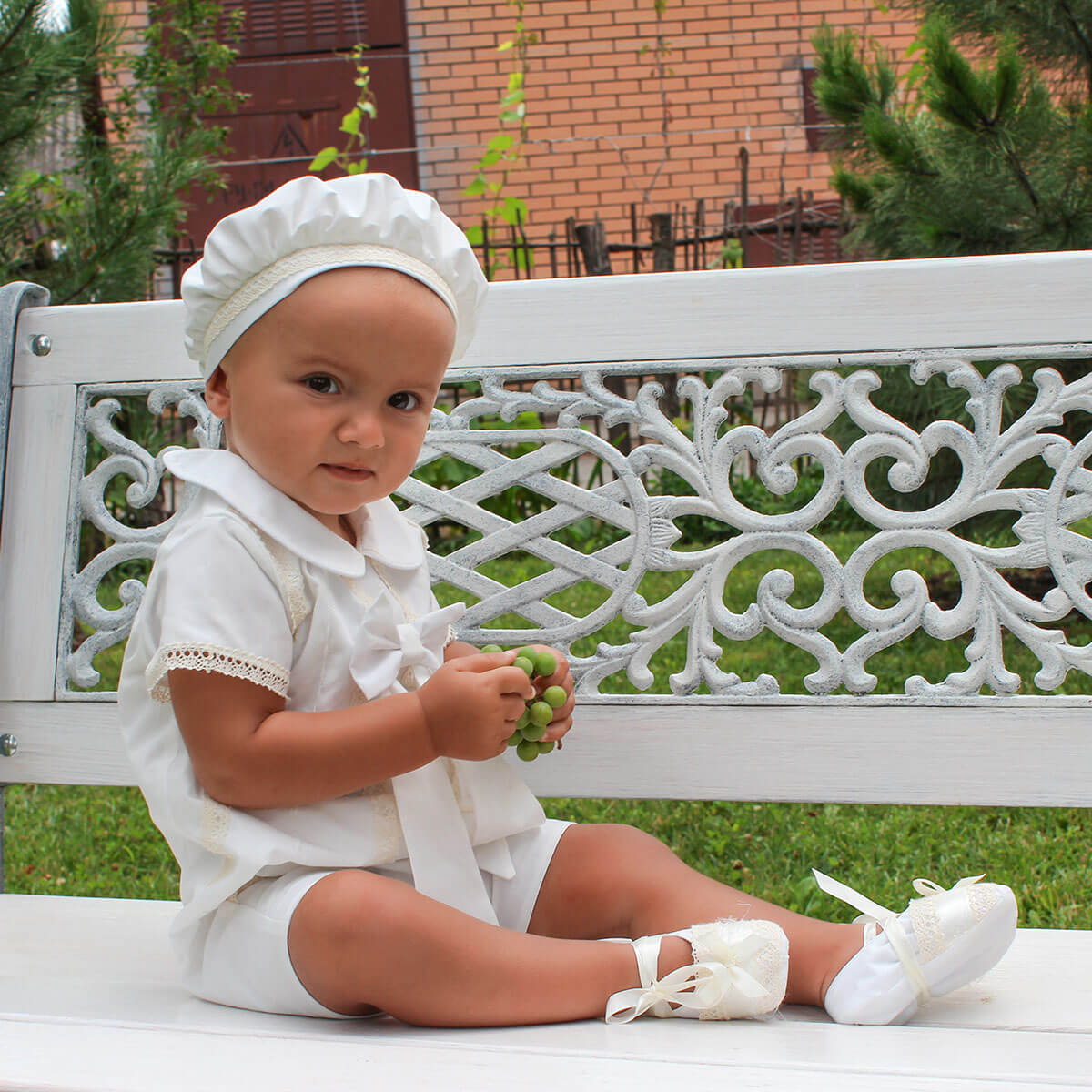 Baby in Boy Christening Hat sitting on a decorative bench outdoors
