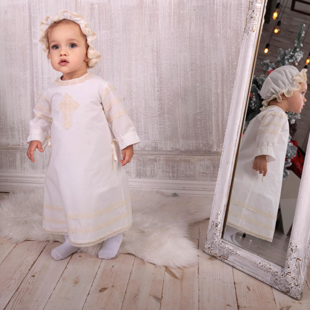 Baby in a Baptism Gown standing next to a mirror with a decorative frame.