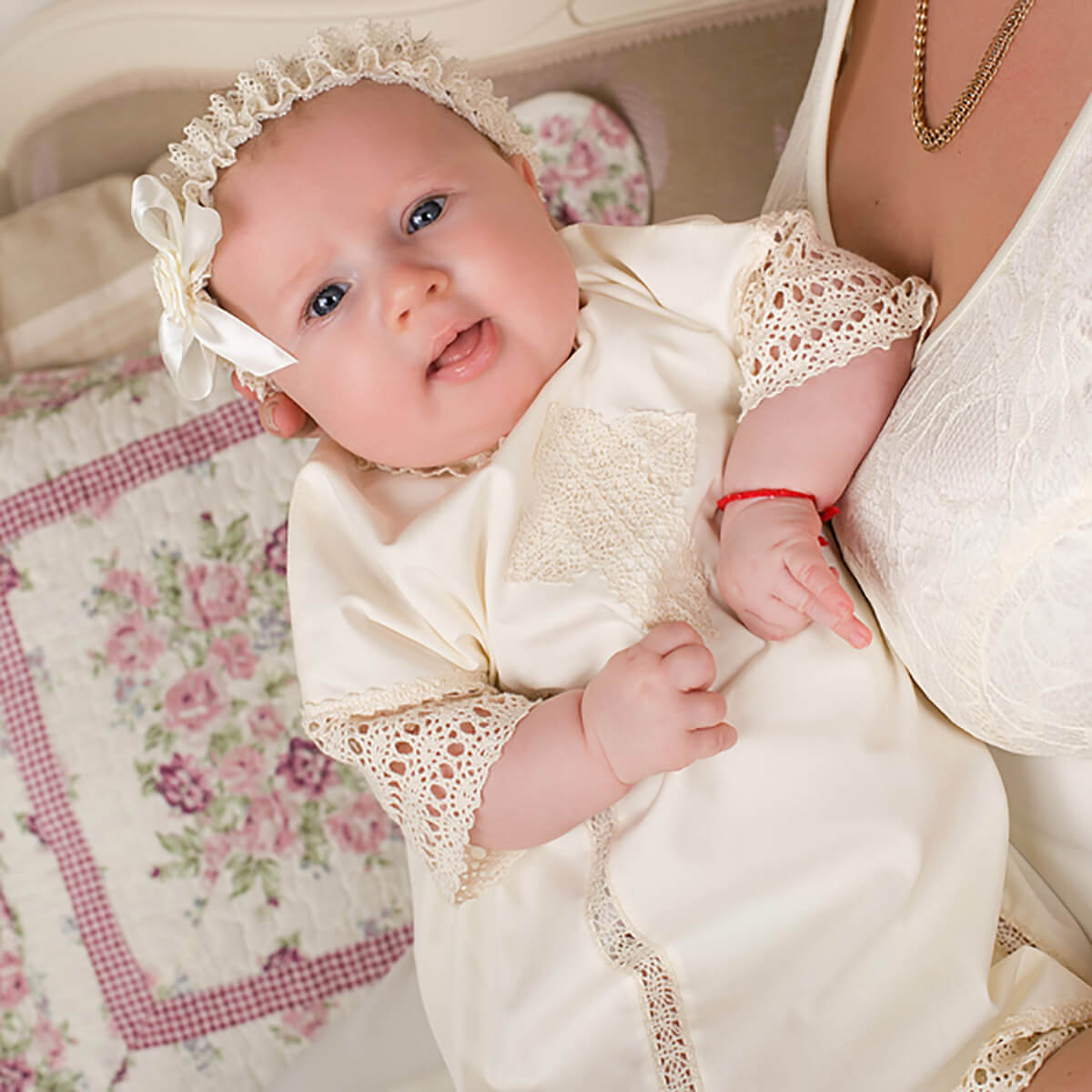 Baby in a white outfit with baby headband held by an adult, sitting on a floral-patterned blanket.