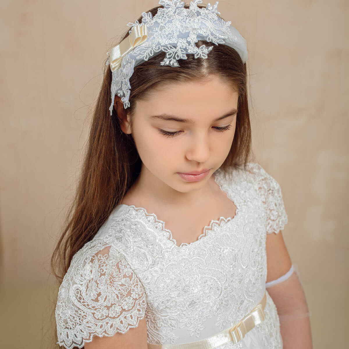 Young girl wearing a white communion lace dress and headband against a beige background