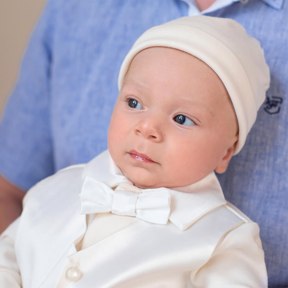 Baby in a Newborn Coming Home Romper with a bow tie and cream hat, standing against a blurred background.