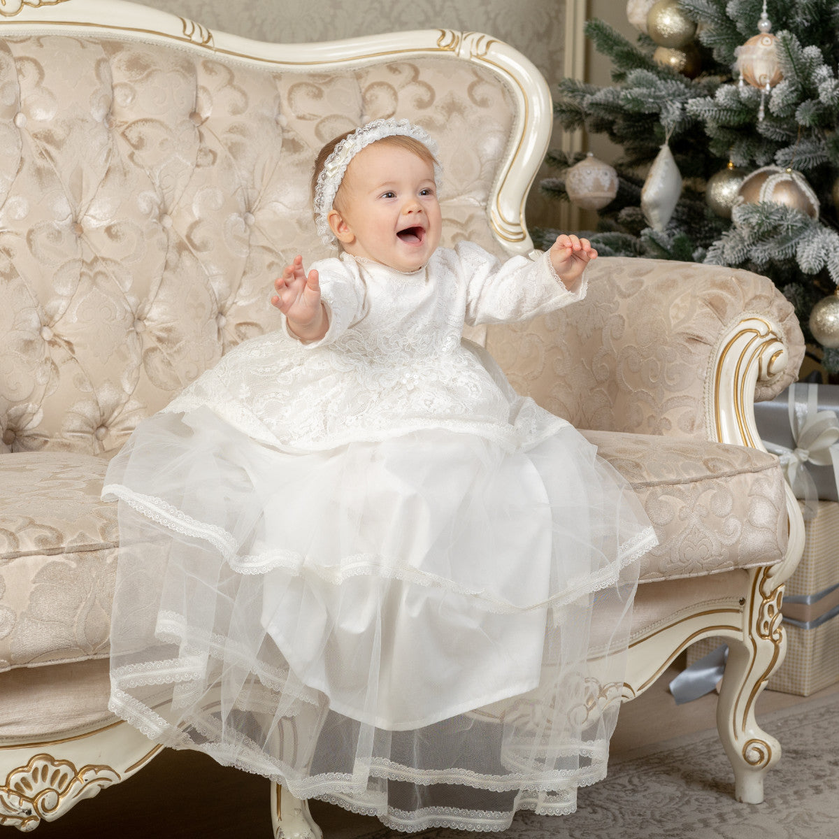 Baby in a Christening Gown for Girls sitting on an ornate chair with a decorated Christmas tree in the background.