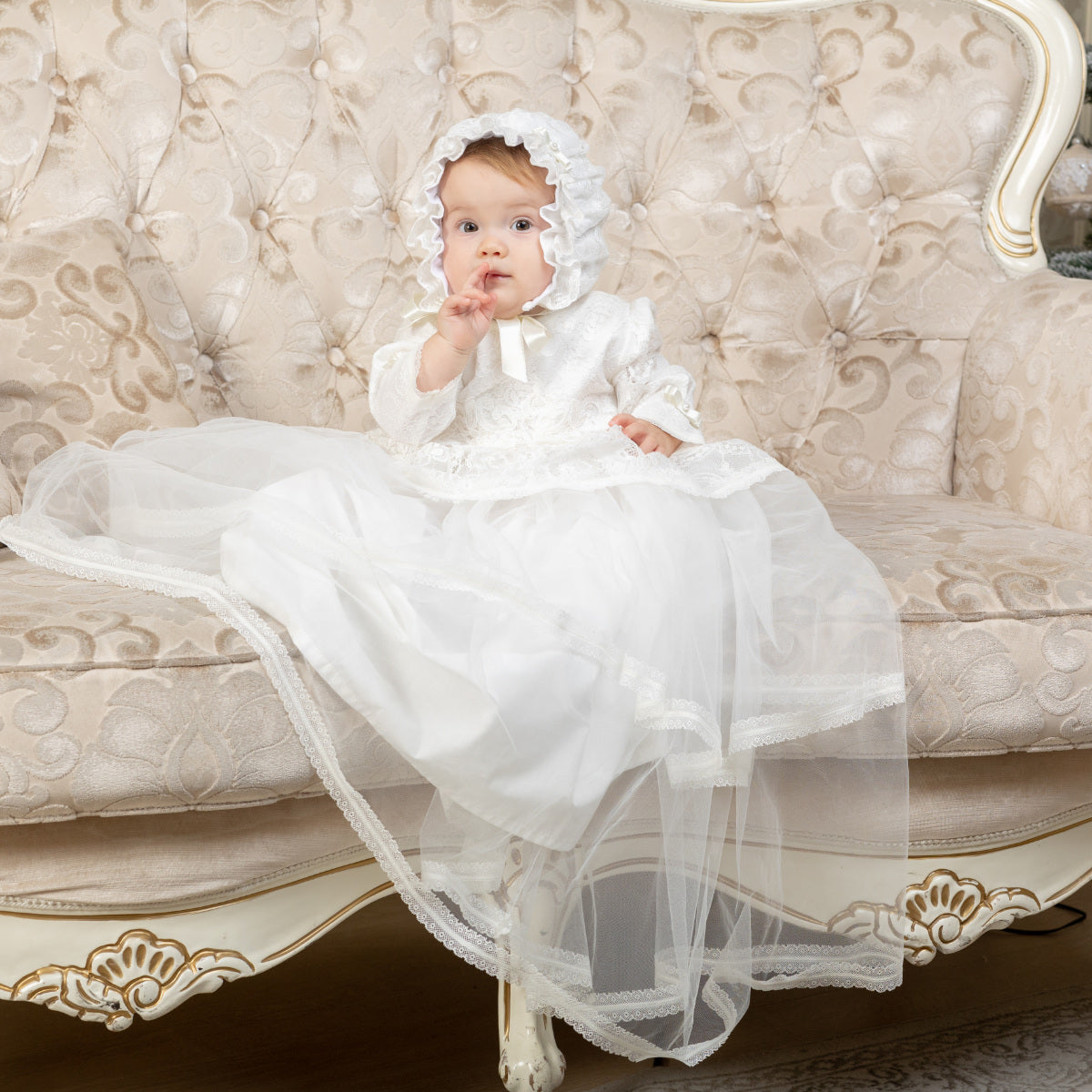 Baby in a Christening Gown for Girls and bonnet sitting on an ornate couch.