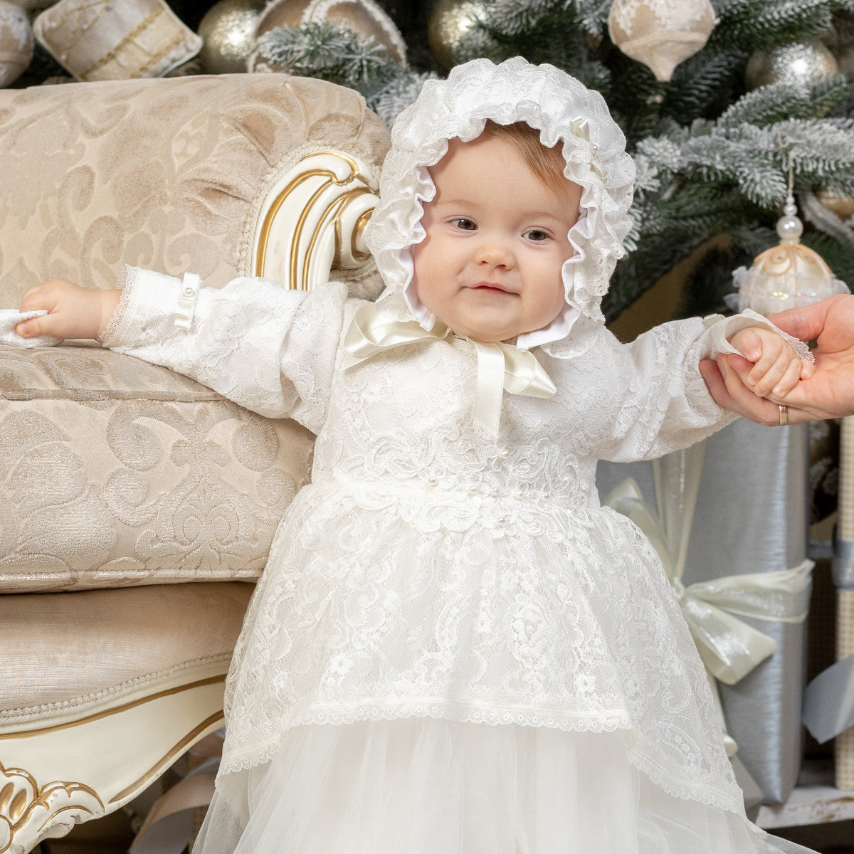 Baby in a white dress and christening bonnet sitting on a decorative chair with Christmas decorations in the background.