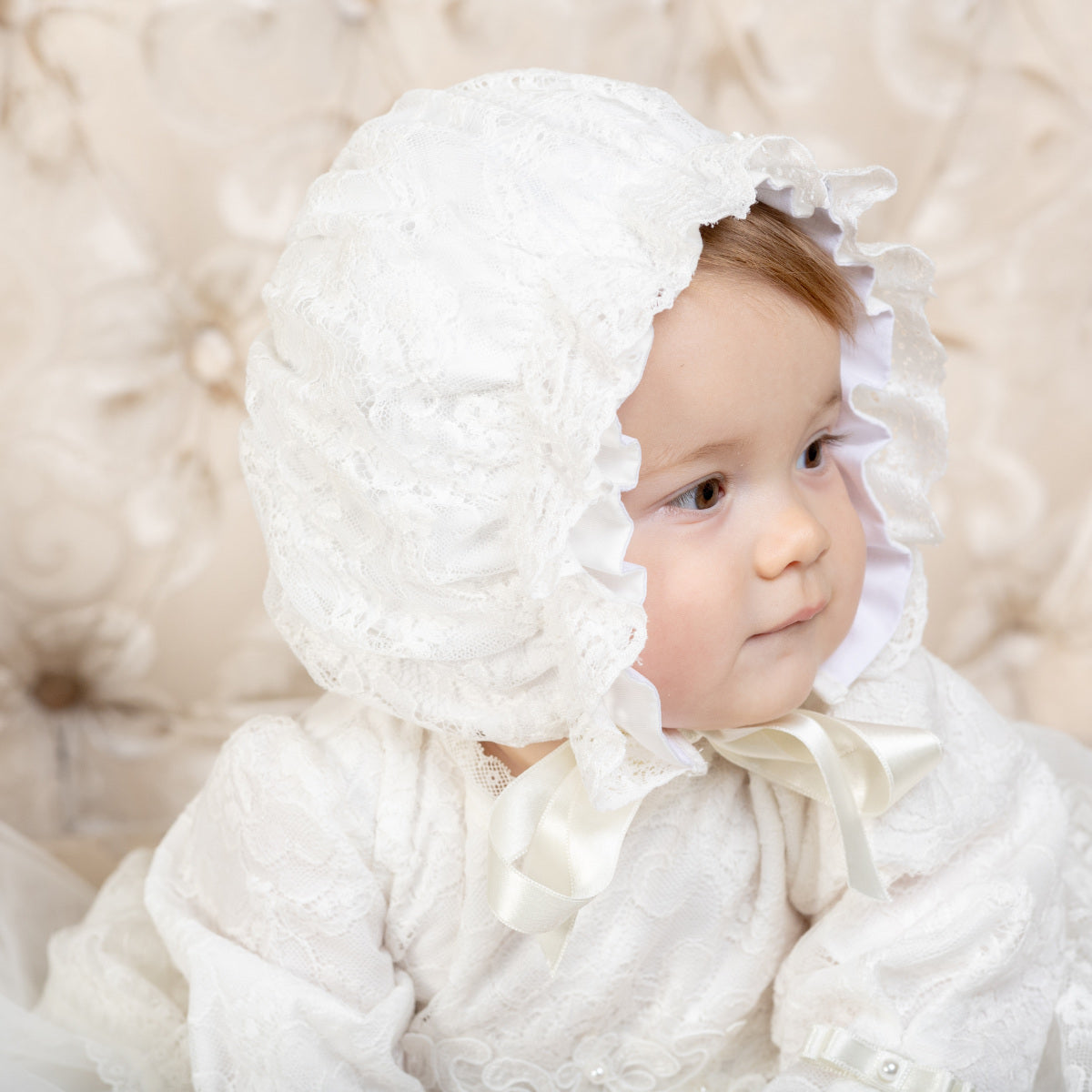 Baby in a lace christening bonnet sitting on a textured beige surface