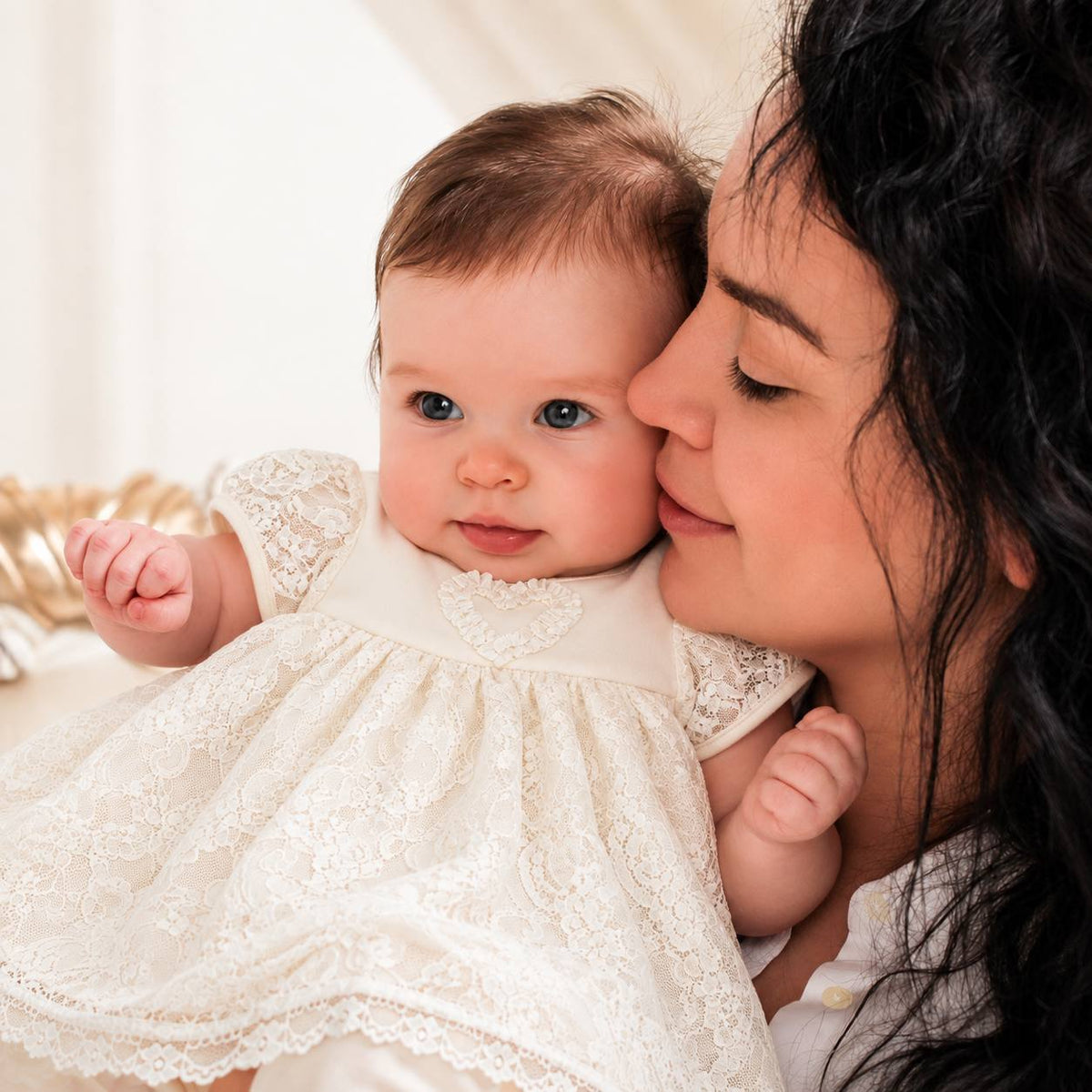 A close-up lifestyle shot of a woman holding and kissing a baby on the cheek. The baby is wearing a cream-colored lace dress.