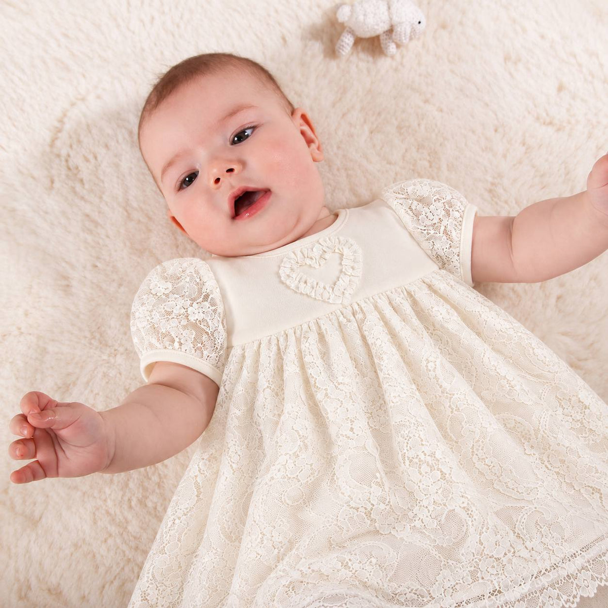 A baby lying on a soft cream faux-fur surface, wearing an ivory lace baby dress with short sleeves and a heart detail on the chest.