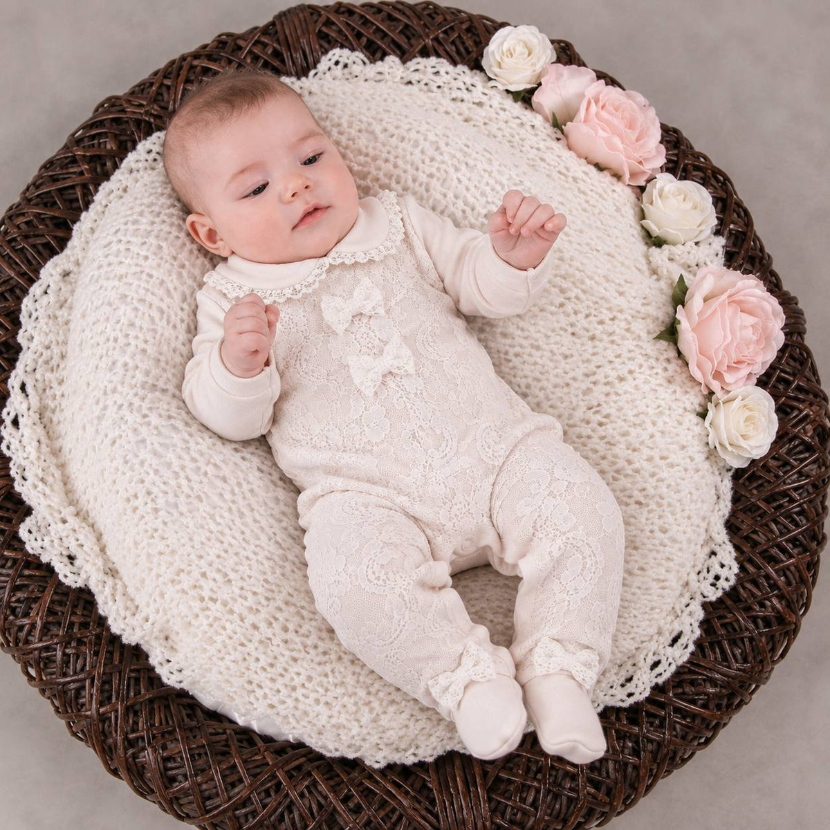 A baby wearing a cream lace footed romper and booties lies on a cream crochet blanket inside a dark brown woven basket, with pink and white faux roses along the edge.