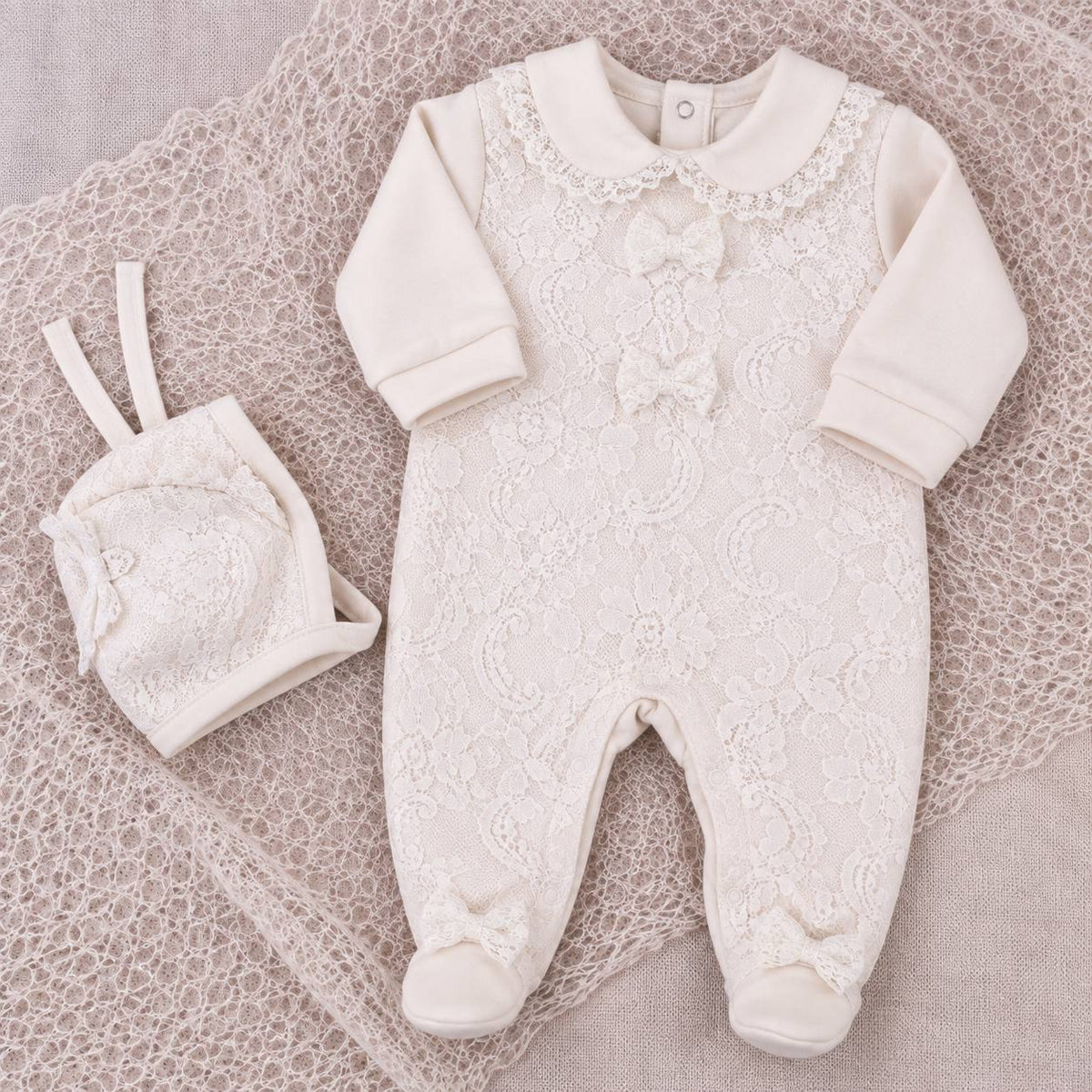 An off-white/cream baby footed one-piece romper with long sleeves, lace floral embroidery, and a bow on the chest, shown flat-laid on a textured lace blanket alongside a matching small hat.