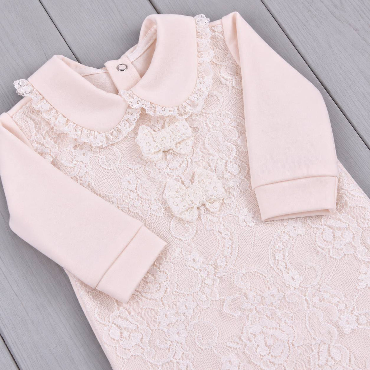A pale pink long-sleeve baby dress with a lace front panel, ruffled Peter Pan collar, and matching fabric bow details, laid flat on a gray wood plank surface.