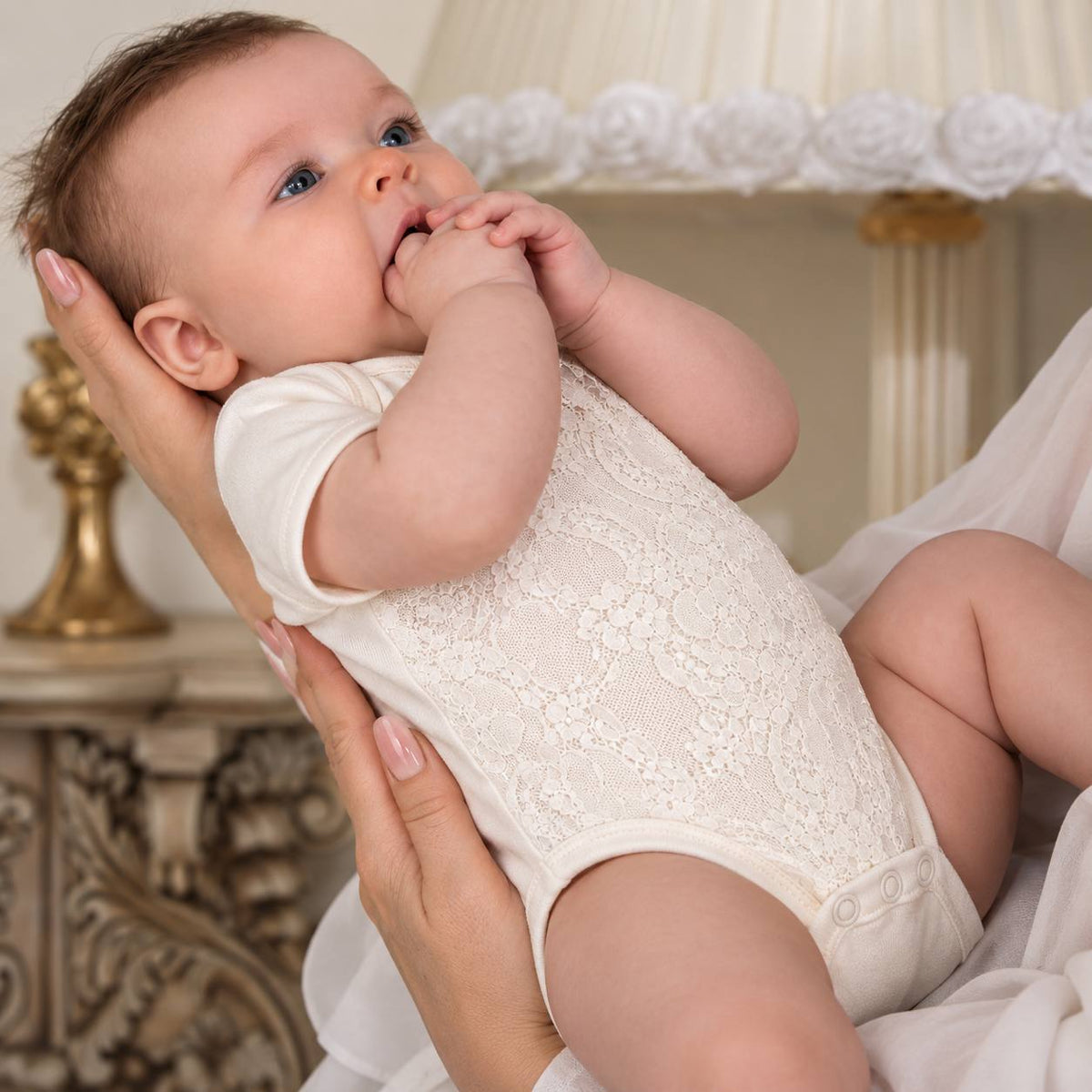 A baby wearing a cream lace bodysuit held by an adult in a softly lit indoor setting with a lamp and ornate furniture in the background.
