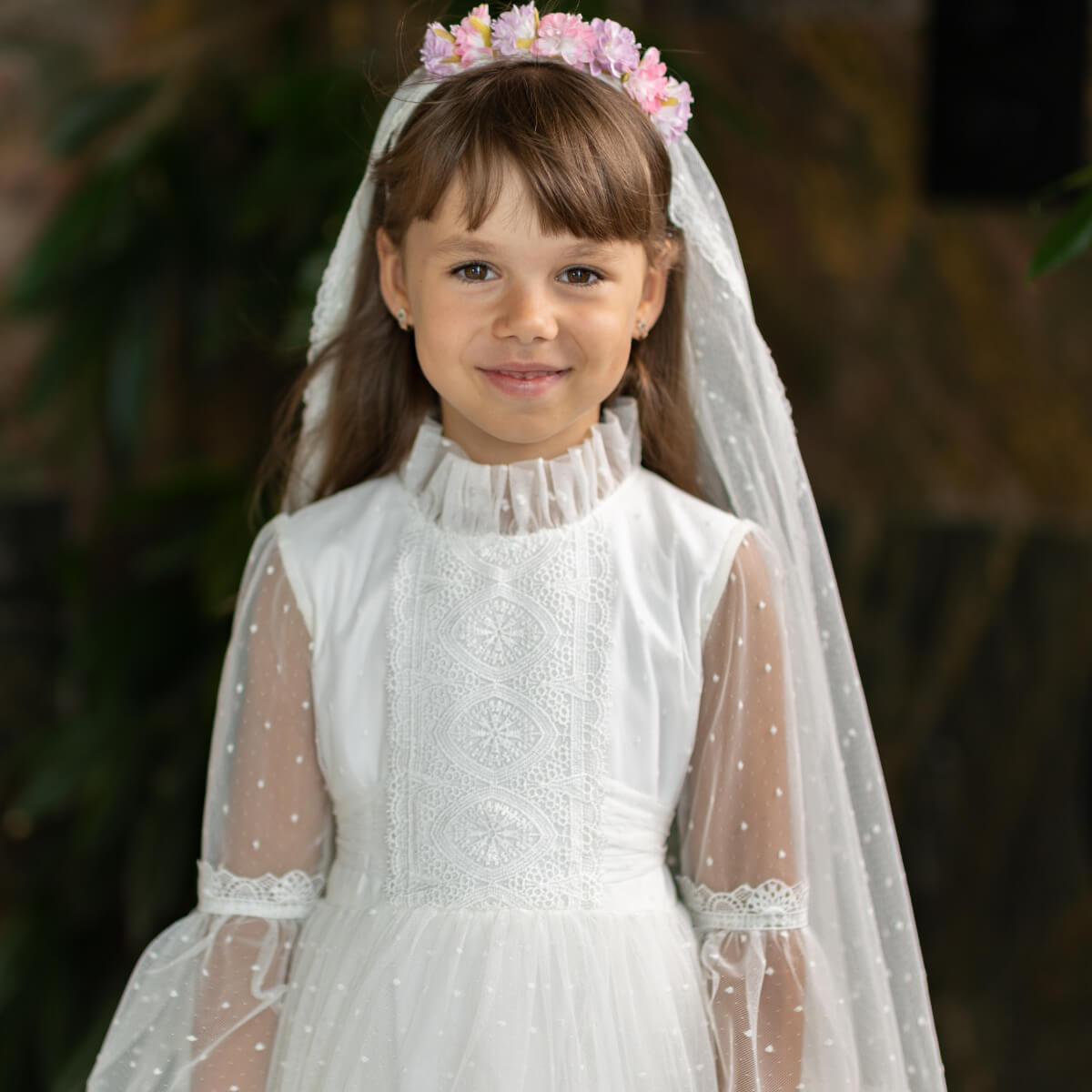 Young girl in a white first communion dress with a veil and floral headband against a blurred natural background