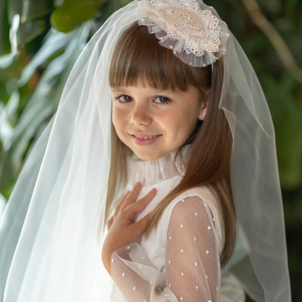 Young girl wearing a white dress with a First Communion Veil and lace headband, standing outdoors.