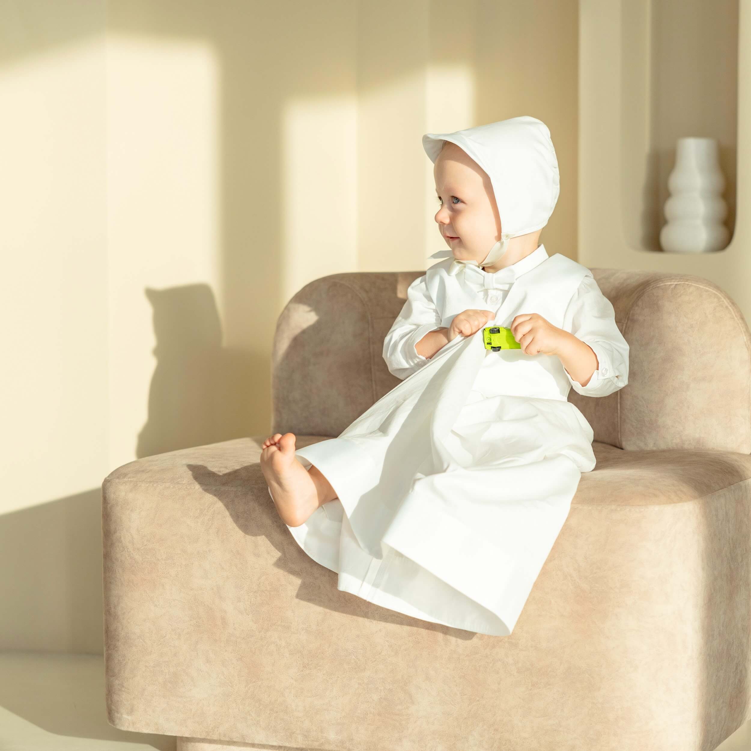 Child in a white outfit sitting on a beige couch holding a green toy.