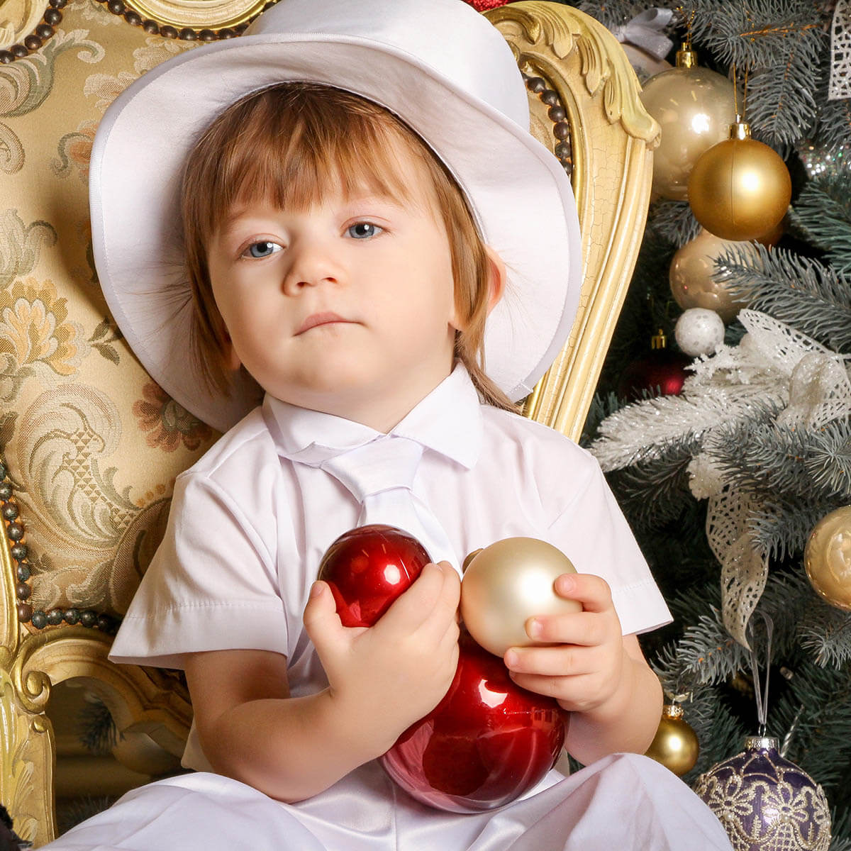 Child in a white outfit and Boy Christening Hat holding Christmas ornaments in front of a decorated tree.