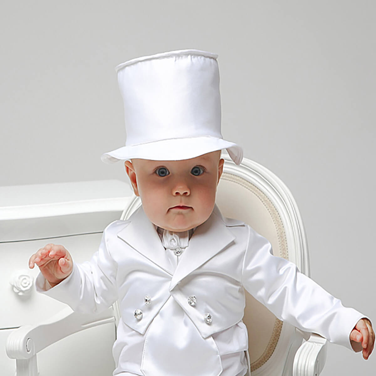 Baby in a formal white outfit with a Boy Christening Hat sitting on a white chair.