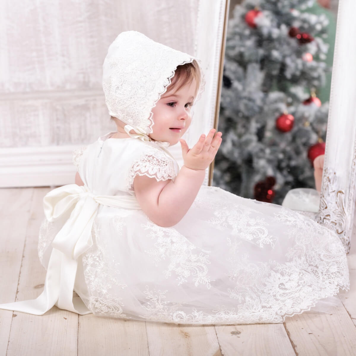 Child in a Elegant Christening Dress and bonnet sitting in front of a decorated Christmas tree.
