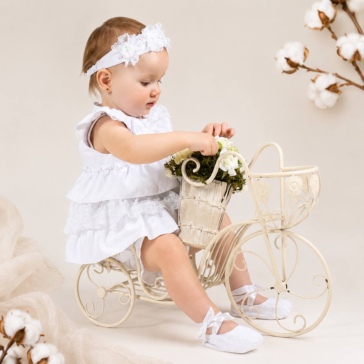 Baby in a white dress sitting on a small white stool with a basket of flowers, surrounded by cotton plants.
